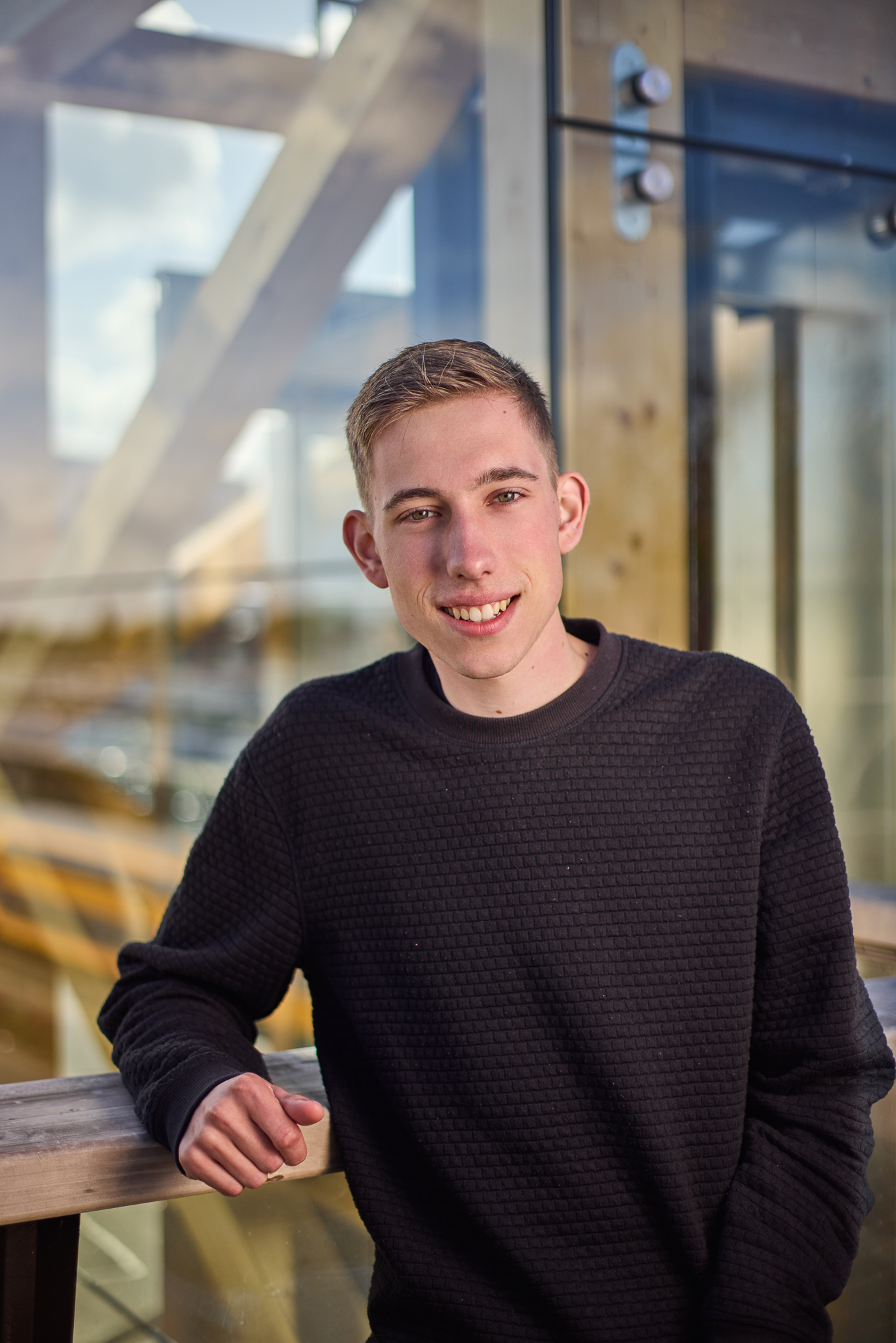 Jongeman in een zwart trui leunt op een houten railing met een glazen muur op de achtergrond.