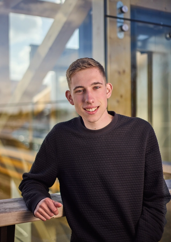 Jongeman in een zwart trui leunt op een houten railing met een glazen muur op de achtergrond.