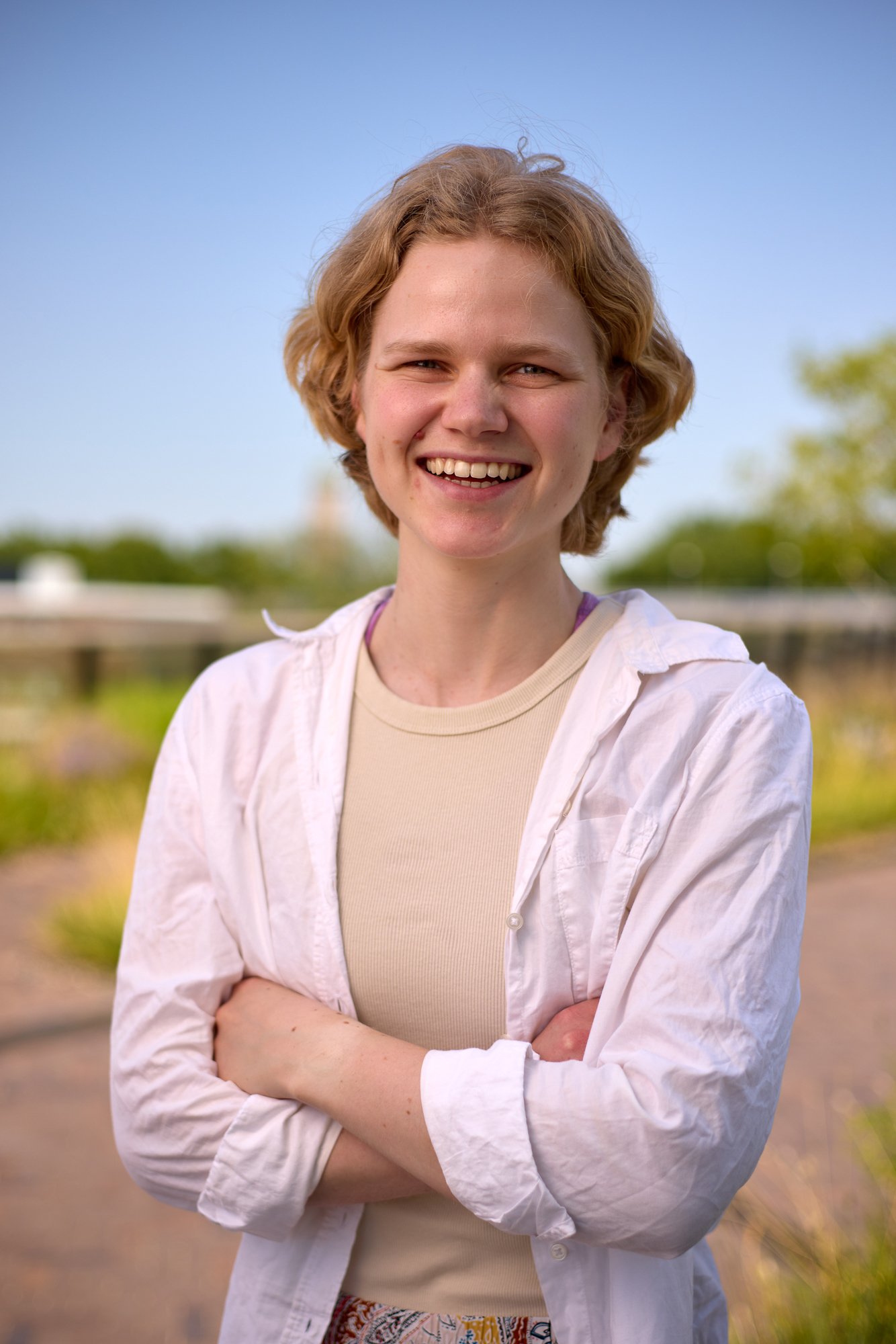 Jonge vrouw met gekruiste armen, staande in een groene omgeving met een blauwe lucht op de achtergrond.