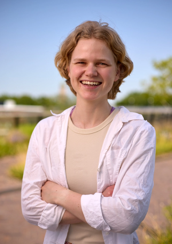 Jonge vrouw met gekruiste armen, staande in een groene omgeving met een blauwe lucht op de achtergrond.