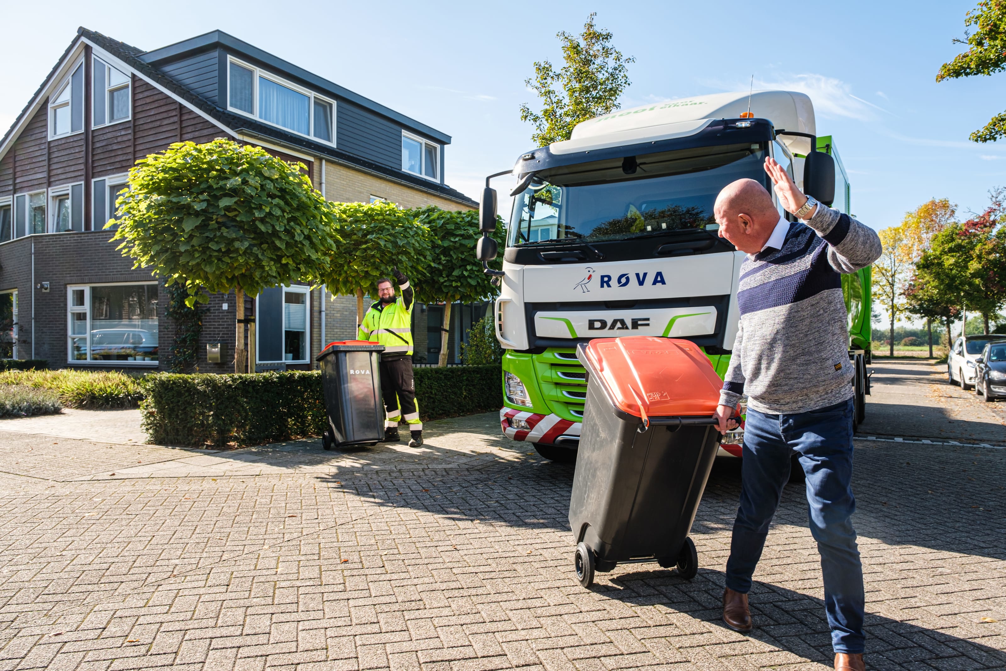 Sfeerfoto van een ROVA medewerker naast een ROVA afvalwagen, zwaaiend naar een inwoner met een groencontainer